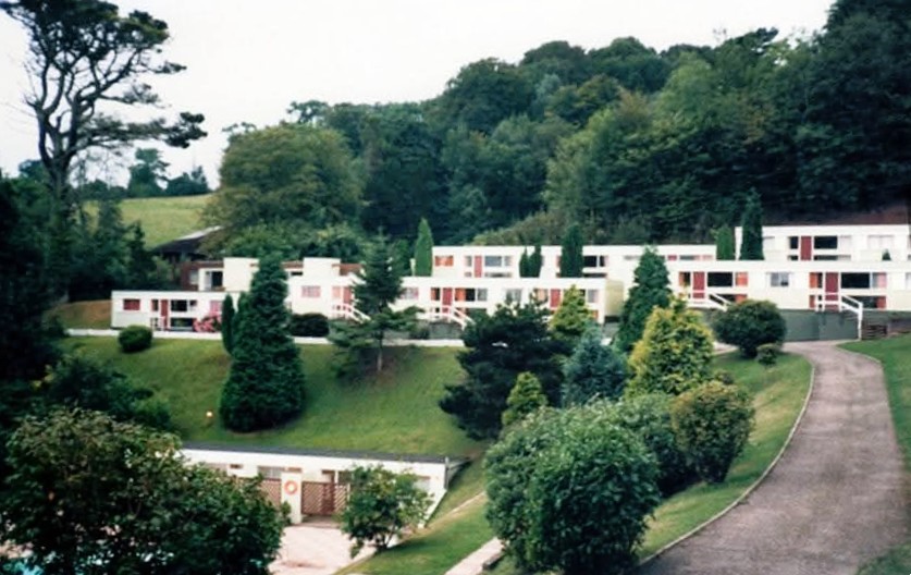 Several blocks of chalets, with three or four in each block. They are single storey but sitting on terraces of a slop with woods behind them and a grassy slope down to a swimming pool in front. The chalets are rendered with, with red or wooden doors and panels.