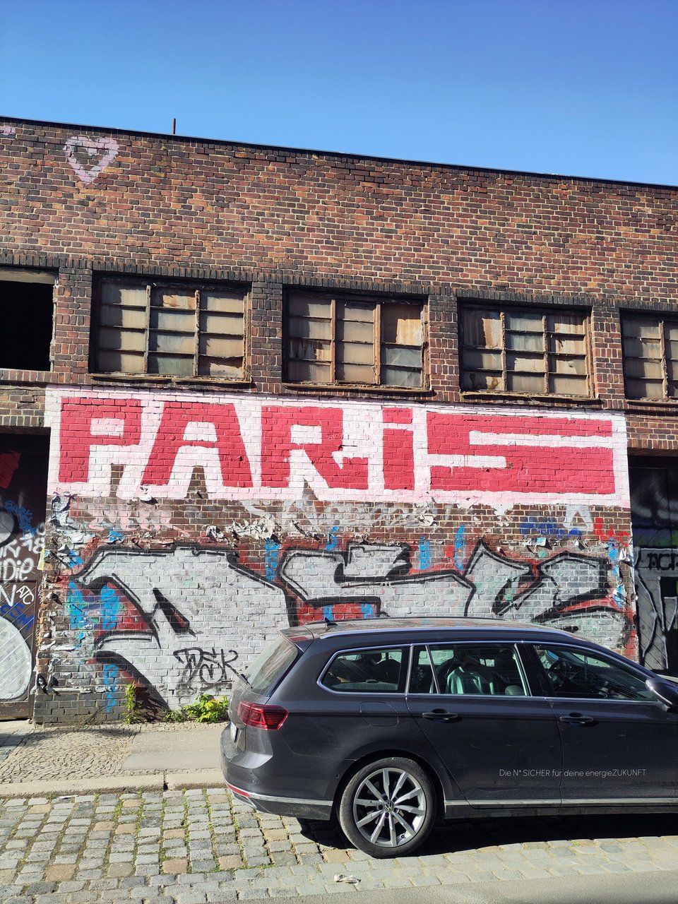 An der Klinkerwand eines alten, anscheinend leerstehenden Fabrikgebäudes steht in großen Graffiti-Buchstaben "Paris". Davor steht ein PKW-Kombi.