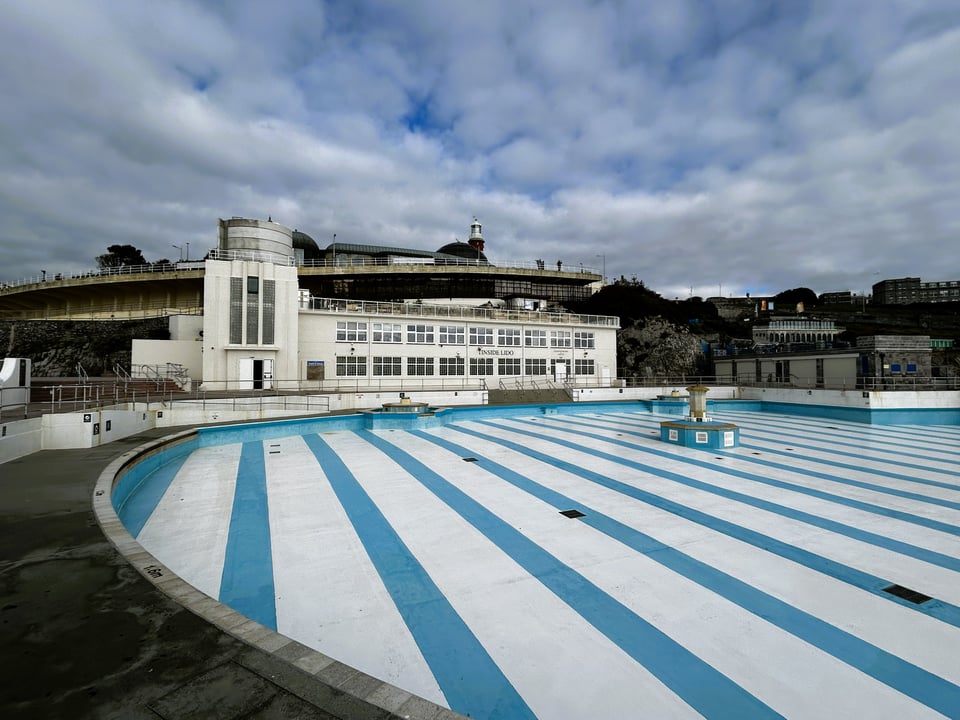 A large empty swimming pool with two tone blue stripes on the bottom, in front of a large white Deco building. It has a three storey staircase to the left, and a two storey long block with windows to the right.