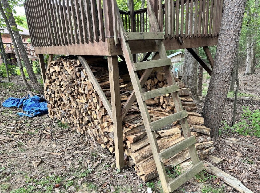 An orderly pile of fire wood underneath the steps of a treehouse.