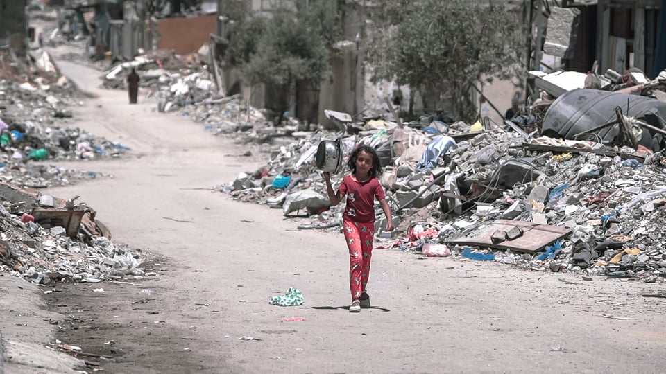 A girl in a red Mickey Mouse shirt and red pants carries an empty pot down a street lined on each side with rubble.