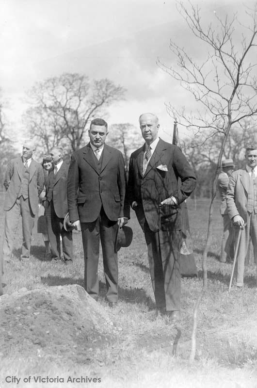 Black and white photo. Two men stand around a pile of dirt and a tree.