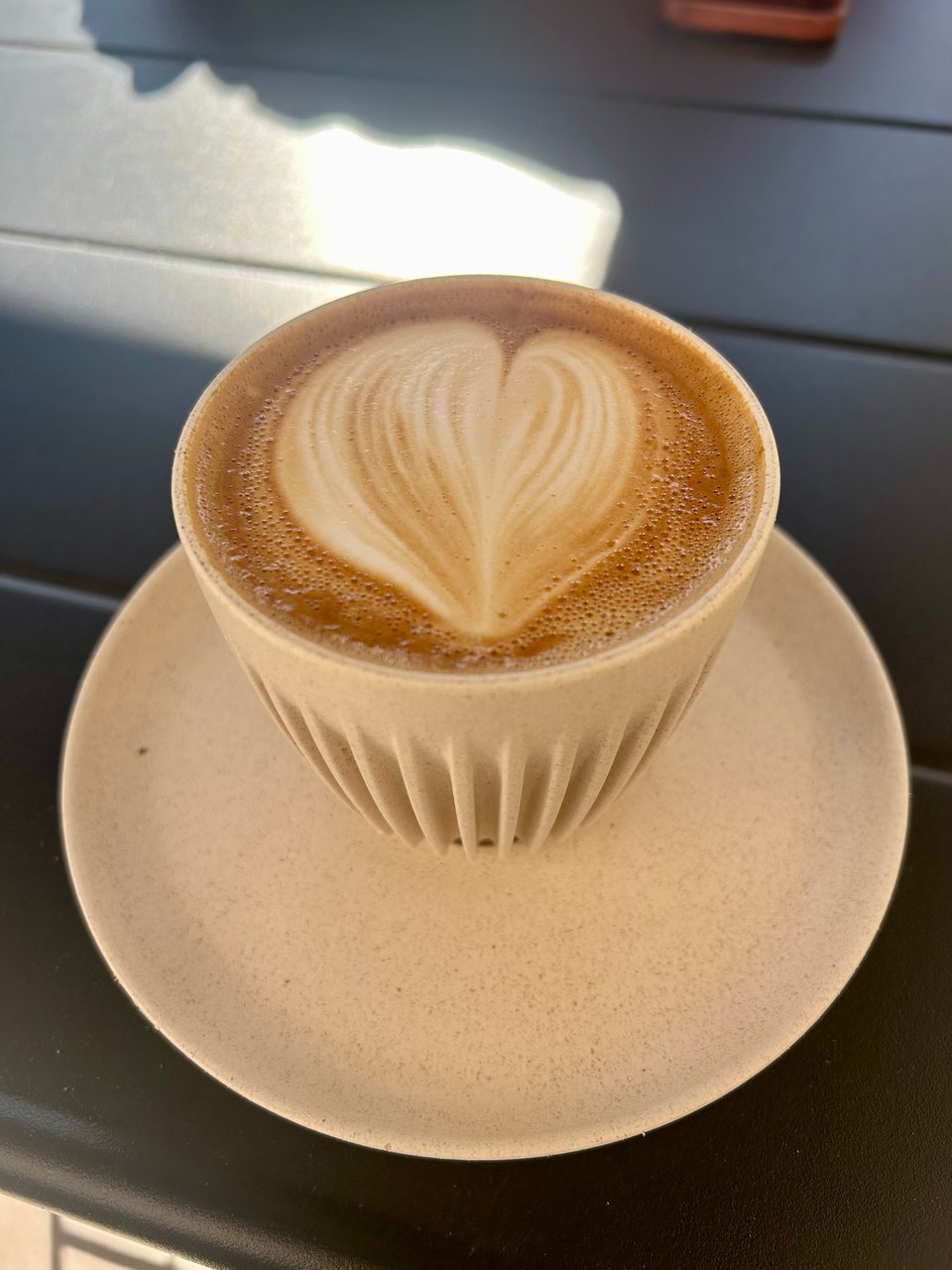 A flat white coffee with latte art in a cream ribbed cup on a cream saucer, which is on a dark grey table. Image by Rowan Ambrose.