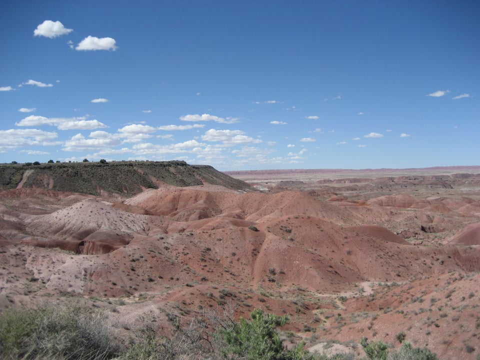 Open badlands, the soil is deep red in color and the land is crinkled. There are some shrubs in the foreground.