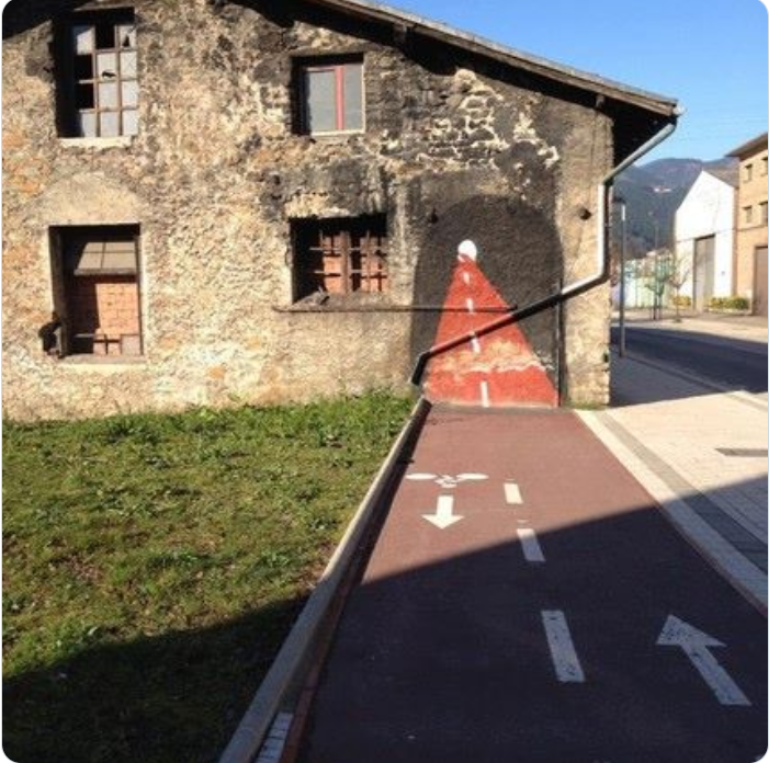 Image of a separated bike lane that ends abruptly against the wall of a house. Someone has painted a cartoon-style continuation of the bike lane, inviting you to cycle straight into the wall