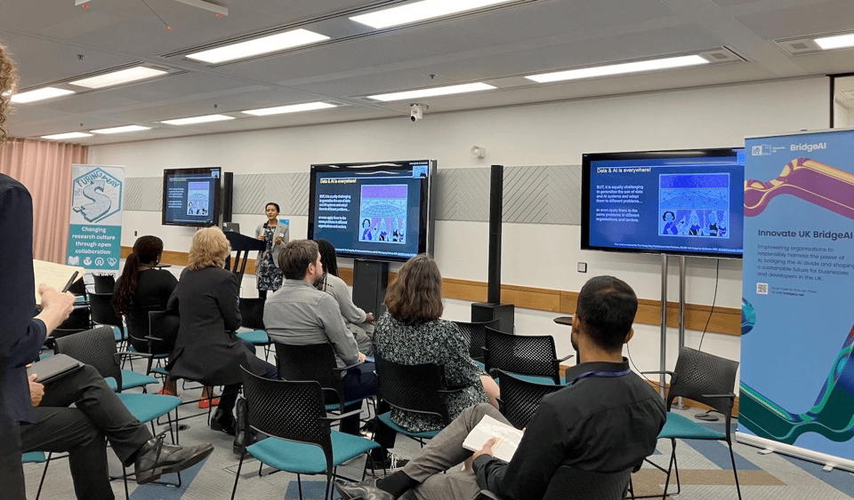 Alt: A picture from the event with Malvika speaking to the attendees with one Turing Way banner on her right and BridgeAI banner on her left.