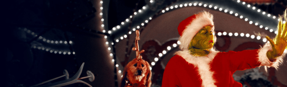 The grinch, an unexplained green entity, is dressed as santa claus. A dog with a raindeer horn strapped to his head sits beside him.