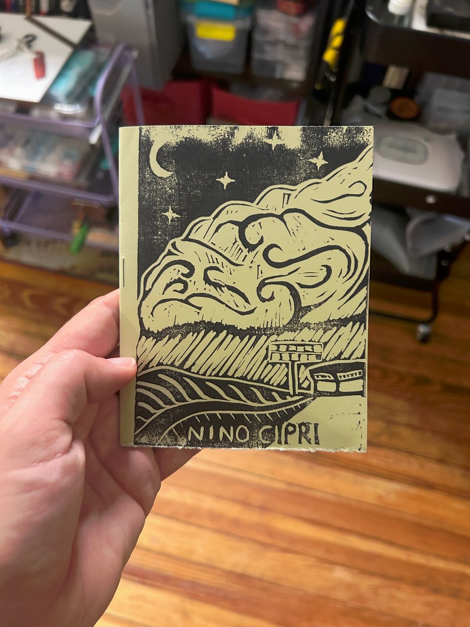 Nino holds a small booklet in their hand, the cover of which has a linoprint of a brightly lit waffle house beneath a giant storm cloud.