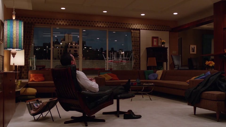 A man in a suit relaxes in a modern mid-century living room, featuring a large sectional sofa, colorful cushions, and a cityscape view at dusk.