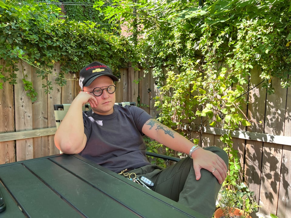 a white bald masc with black circular glasses and a black cap sits at an outdoor table with their hand resting on their face and another hand on their knee, crossed over their other leg