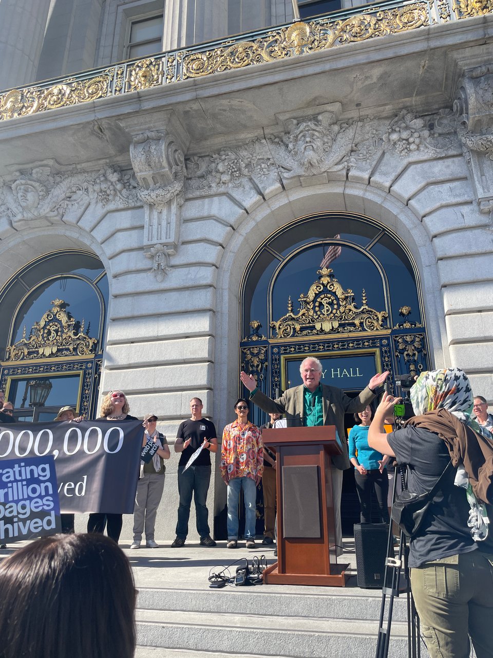 A man talks at a podium in front of City Hall surrounded by people clapping and holding signs.