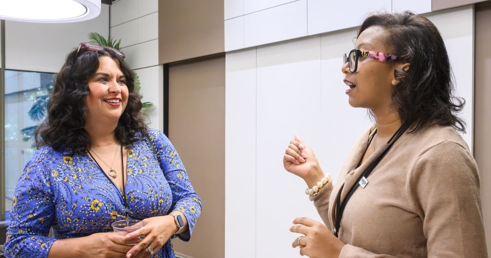 Two women are having a lively conversation indoors, with one gesturing animatedly while the other listens attentively.