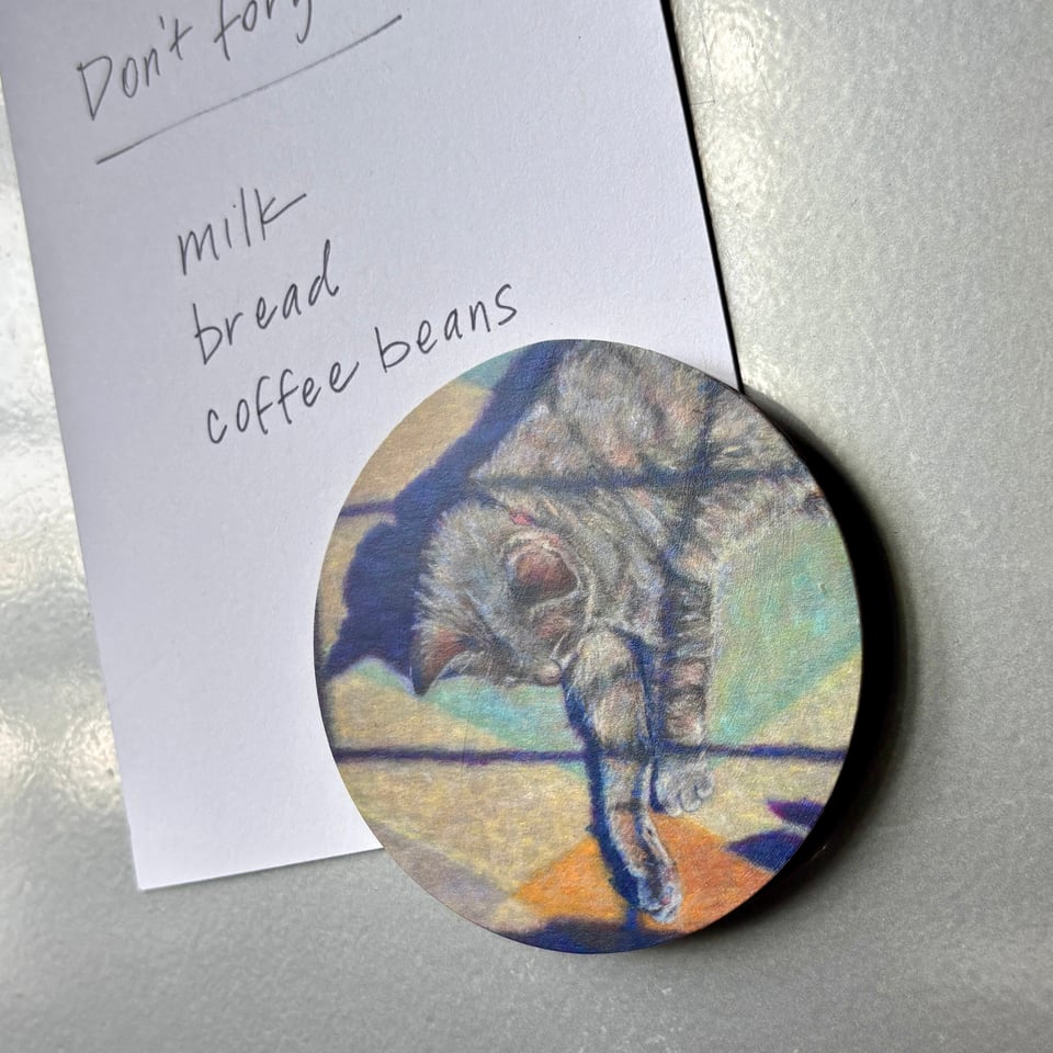 round magnet with a painting of a cat holds a list of groceries on a fridge