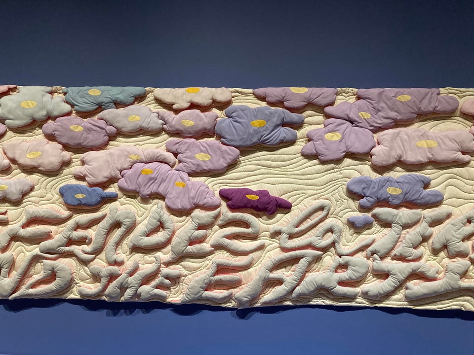 A large quilt with multi-coloured flowers on top, and defendez la terre, defend the earth in bubble letters below