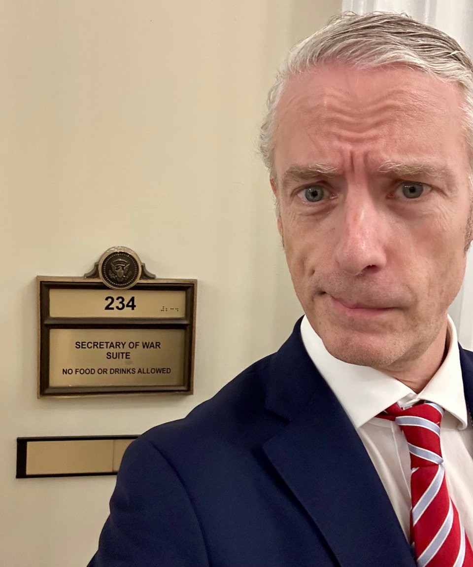 Selfie of white man with nervous look on his face, in blue suit, white dress shirt and red/light blue tie standing in front of of a door sign that reads "SECRETARY OF WAR SUITE"