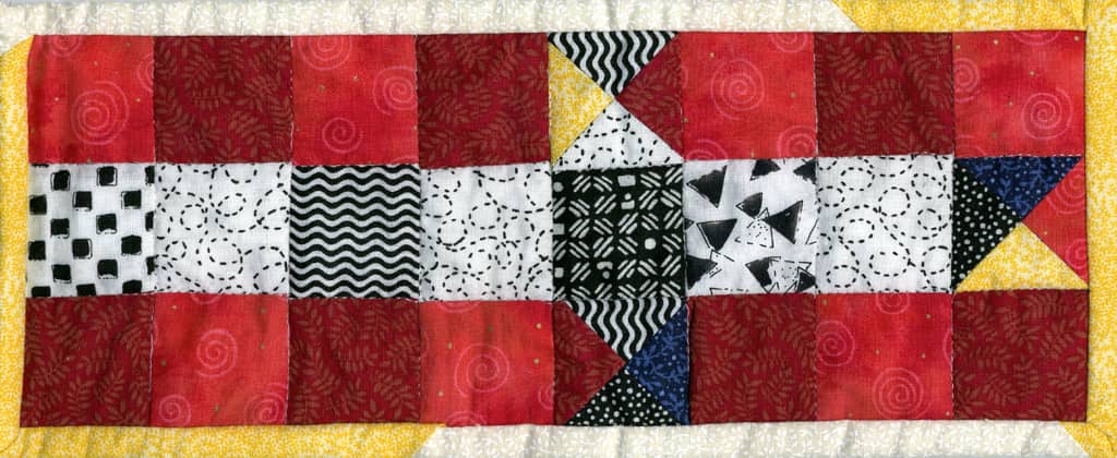 Photo of one arm of a Pacheesi game board that is also a patchwork quilt. There are fabrics in red, yellow, and black & white.