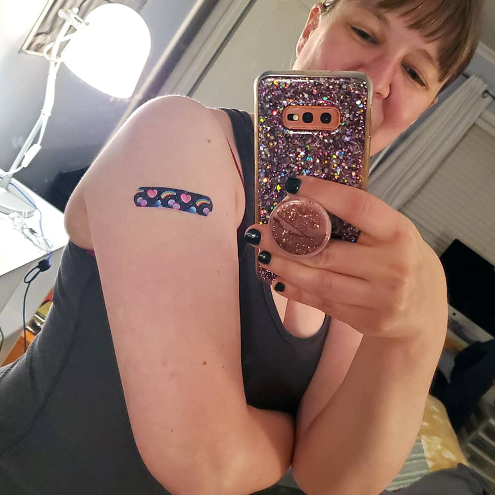 Mirror selfie of Sarah in her home sporting a purple bandaid that includes pink hearts and rainbows.