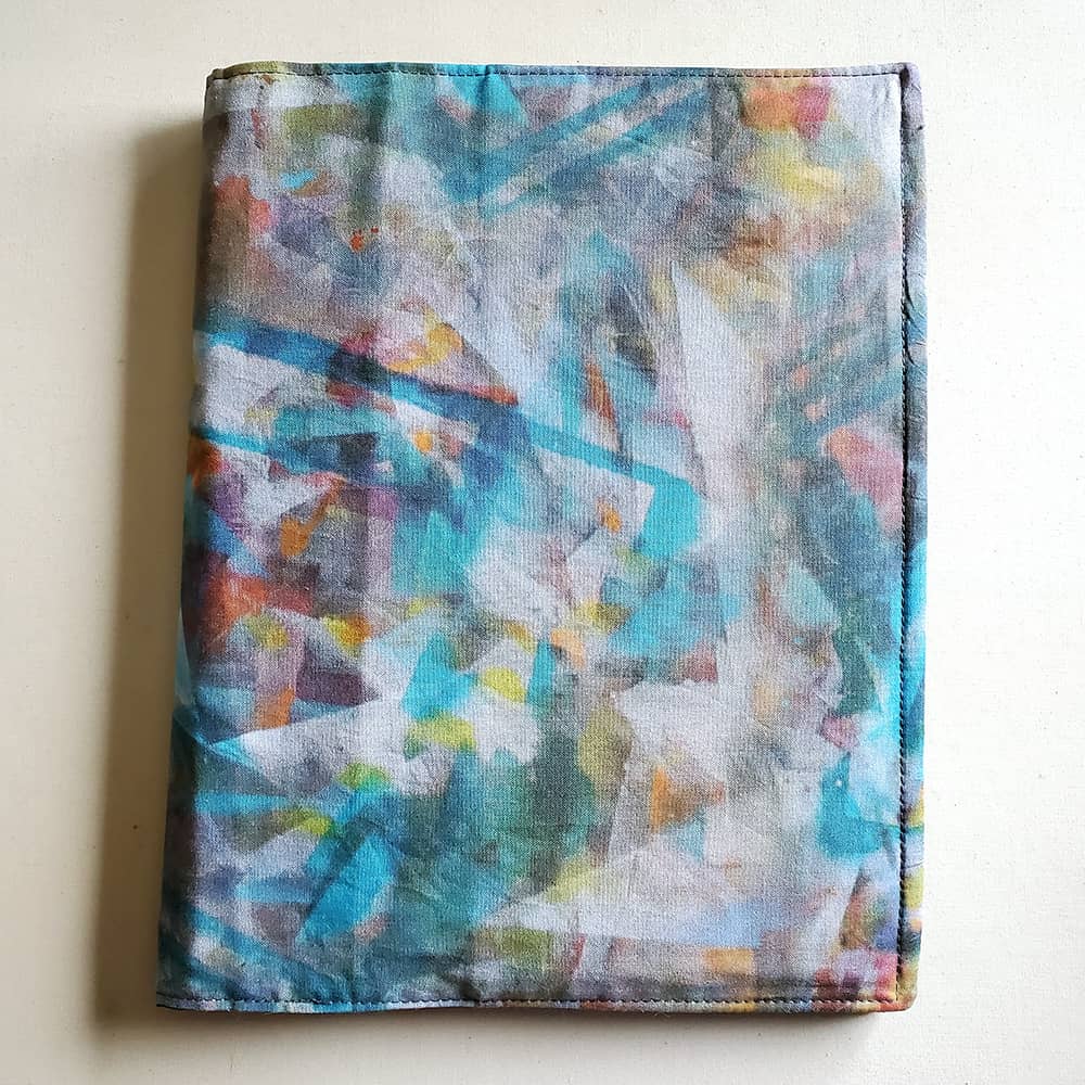 Photo of a fabric notebook cover. The image printed on the fabric is of a tabletop with dried paint all over it.