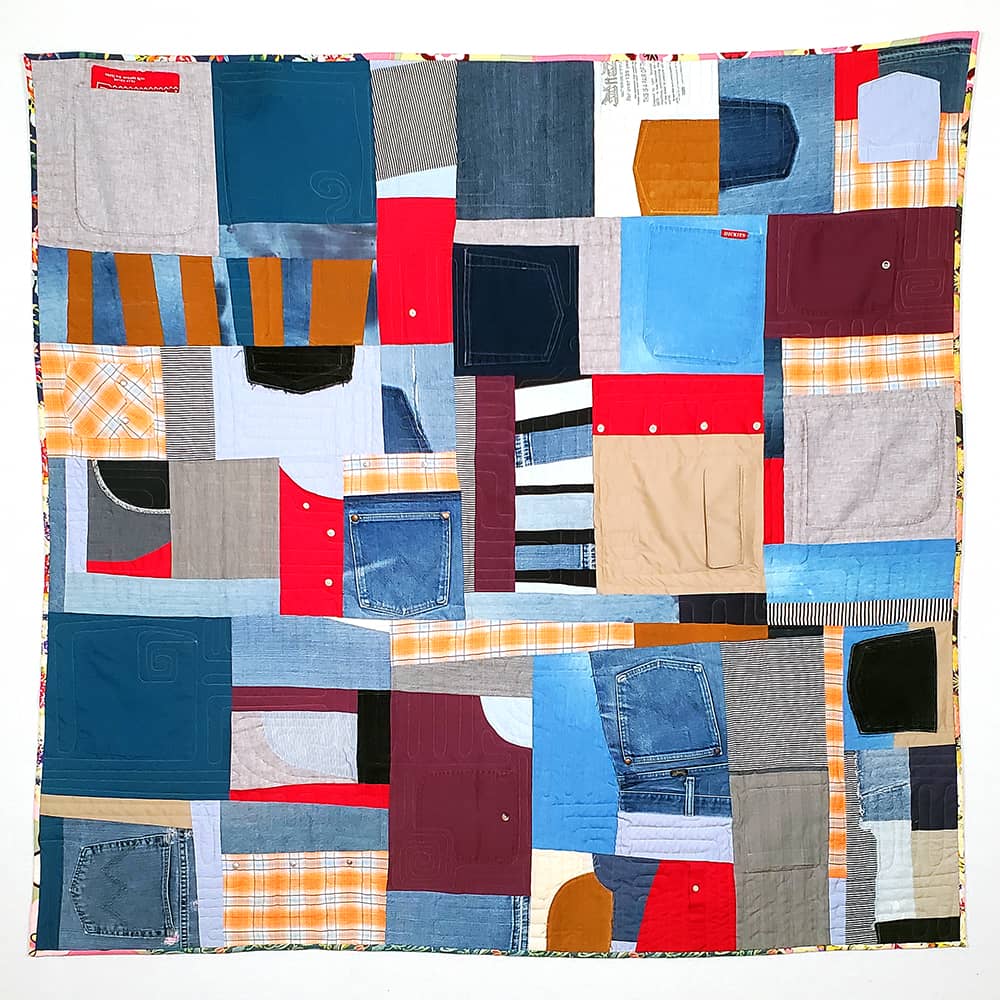 Patchwork quilt made from upcycled garment fabrics, including many pockets. The fabric colors include red, blue, beige, grey, gold, and maroon.