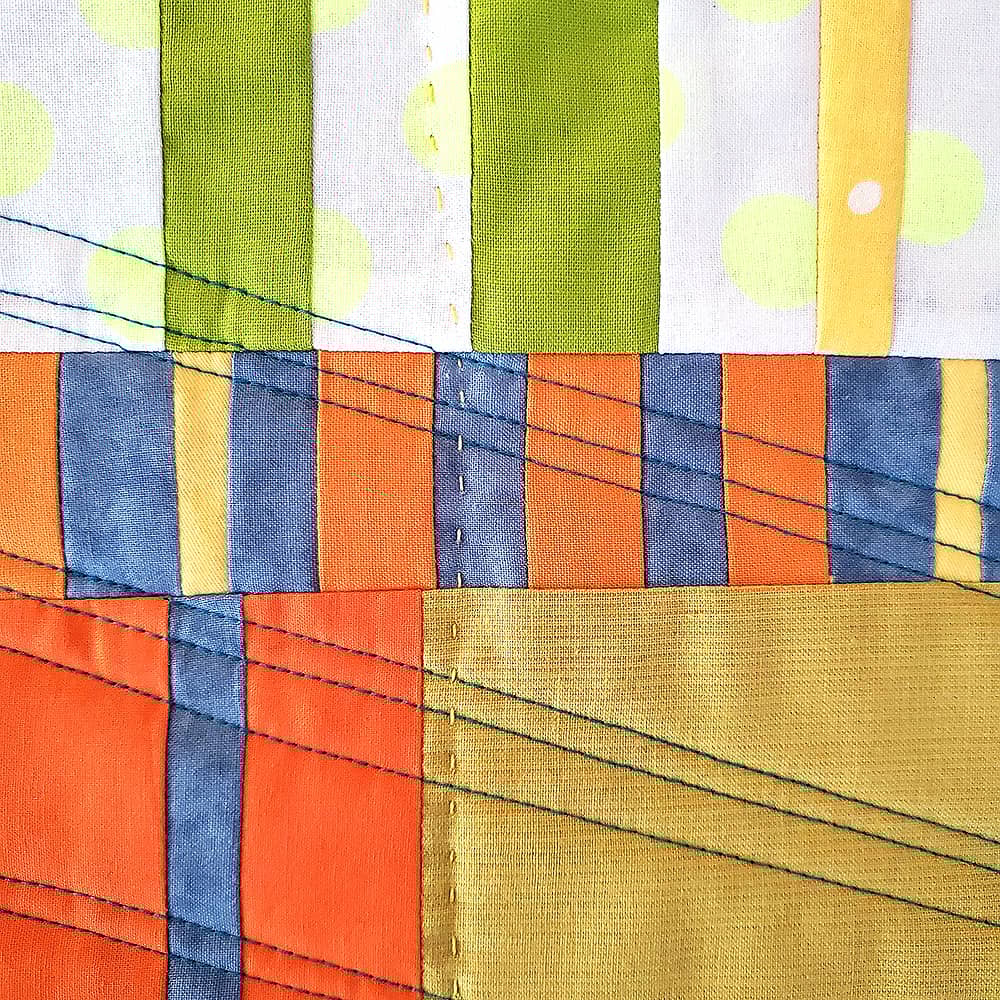 Detail photo of a small patchwork quilt with fabrics in orange, yellow, white, light green, and gray. There are diagonal rows of stitching in dark thread.